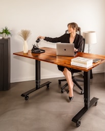 A woman in a black suit is seated at a wooden desk with an open laptop in front of her. She is reaching for a traditional black rotary phone on the desk. The workspace includes decorative elements such as a vase with dried grass, a small plant, and a stack of books. A lamp stands in the background.