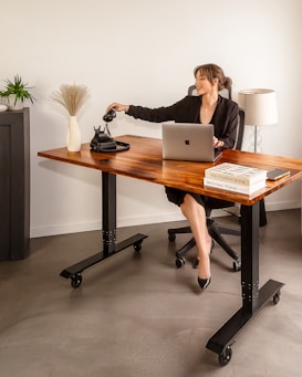 A woman in a black suit is seated at a wooden desk with an open laptop in front of her. She is reaching for a traditional black rotary phone on the desk. The workspace includes decorative elements such as a vase with dried grass, a small plant, and a stack of books. A lamp stands in the background.