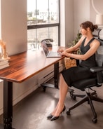 A woman sits in a modern office setting at a large wooden desk, working on a laptop. She is dressed in a black dress and high heels. The room is well-lit with natural light coming through a large window. An old-fashioned rotary phone and books are on the desk, and a potted plant is placed nearby.