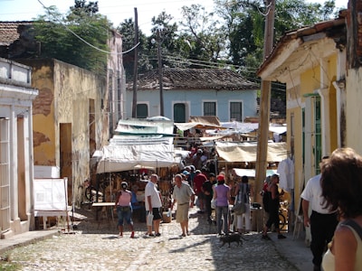 A bustling outdoor market scene on a cobblestone street, surrounded by rustic buildings with peeling paint. Canopies and stalls line the street, filled with various items for sale. Numerous people are browsing the market, some engaged in conversation, while a small dog roams around.
