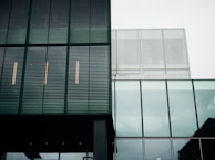 A modern architectural facade with a combination of metal louver panels and large glass sections. The structure features clean lines and a minimalist design. The reflection of the sky is visible in the glass, and the building's geometric patterns create a sense of order.