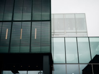 A modern architectural facade with a combination of metal louver panels and large glass sections. The structure features clean lines and a minimalist design. The reflection of the sky is visible in the glass, and the building's geometric patterns create a sense of order.