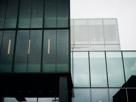 A modern architectural facade with a combination of metal louver panels and large glass sections. The structure features clean lines and a minimalist design. The reflection of the sky is visible in the glass, and the building's geometric patterns create a sense of order.