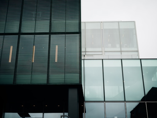 A modern architectural facade with a combination of metal louver panels and large glass sections. The structure features clean lines and a minimalist design. The reflection of the sky is visible in the glass, and the building's geometric patterns create a sense of order.