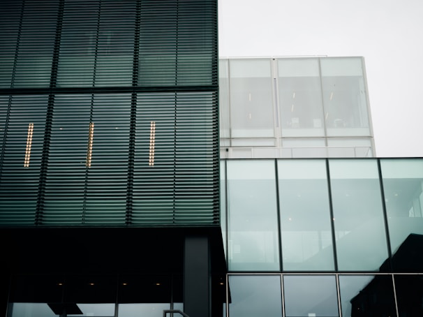 A modern architectural facade with a combination of metal louver panels and large glass sections. The structure features clean lines and a minimalist design. The reflection of the sky is visible in the glass, and the building's geometric patterns create a sense of order.