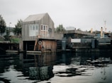 A waterside scene featuring a rustic, wooden building with large windows next to a body of water. The structure is elevated on a platform with a small ladder leading to a dock. Surrounding the building are industrial elements, including another structure with a flat roof. Trees can be seen in the background under an overcast sky, and the water reflects the muted colors of the environment.