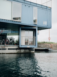 A modern structure made from shipping containers is positioned by the waterfront, with large windows and bicycles visible through the glass. The containers are painted blue and are stacked in a creative architectural design. The water in the foreground has gentle ripples, and the sky is overcast.