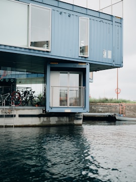A modern structure made from shipping containers is positioned by the waterfront, with large windows and bicycles visible through the glass. The containers are painted blue and are stacked in a creative architectural design. The water in the foreground has gentle ripples, and the sky is overcast.