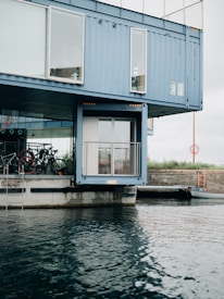 A modern structure made from shipping containers is positioned by the waterfront, with large windows and bicycles visible through the glass. The containers are painted blue and are stacked in a creative architectural design. The water in the foreground has gentle ripples, and the sky is overcast.