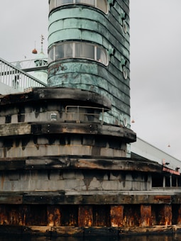 A structure with layered, metallic cladding and visible oxidation on a cylindrical tower. The base is made of concrete with wooden reinforcement layers. Windows are integrated into the upper part, with a weathered appearance throughout.