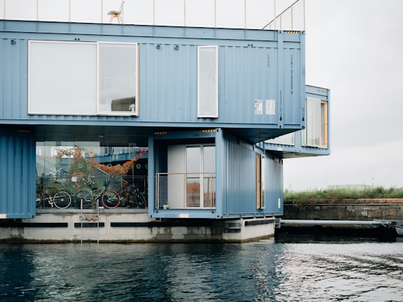 A modern building constructed from stacked blue shipping containers sits by the water's edge. Large windows provide a view of the interior, which includes bicycles and decorated plants. The structure features a minimalist design with an industrial aesthetic, and a lone chair is visible on the rooftop.