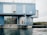 A modern building constructed from stacked blue shipping containers sits by the water's edge. Large windows provide a view of the interior, which includes bicycles and decorated plants. The structure features a minimalist design with an industrial aesthetic, and a lone chair is visible on the rooftop.