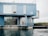 A modern building constructed from stacked blue shipping containers sits by the water's edge. Large windows provide a view of the interior, which includes bicycles and decorated plants. The structure features a minimalist design with an industrial aesthetic, and a lone chair is visible on the rooftop.