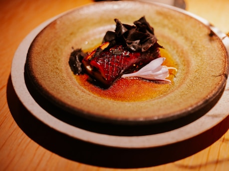 A beautifully plated dish featuring a glistening piece of seared fish atop a rustic ceramic plate. The fish is garnished with dark, leafy greens and a delicate, pink flower petal. The background surfaces are wooden and warmly lit, enhancing the rich colors of the food.