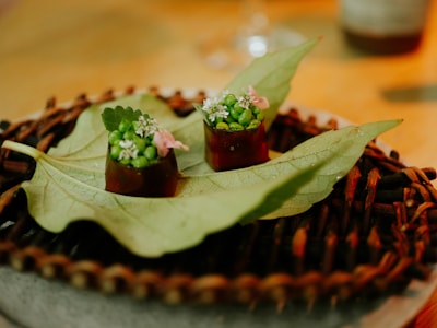 Two small gourmet creations are placed on a large green leaf, which serves as a unique platter. Each creation is garnished with vibrant green peas and delicate white flowers, accompanied by a small pink flower. The items rest atop a woven, rustic mat placed on a circular plate, with a warm, softly lit background enhancing the presentation.