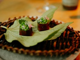 Two small gourmet creations are placed on a large green leaf, which serves as a unique platter. Each creation is garnished with vibrant green peas and delicate white flowers, accompanied by a small pink flower. The items rest atop a woven, rustic mat placed on a circular plate, with a warm, softly lit background enhancing the presentation.