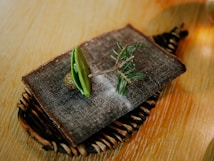A minimalist food presentation consisting of a small green taco-like item resting on a textured fabric atop a wooden board. A sprig of evergreen appears to be part of the garnish, providing a rustic and natural appeal with surrounding elements in soft focus.