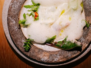 A gourmet dish featuring foamy sauce, delicate greens, and a small yellow flower arranged on a rustic ceramic plate. The presentation is artistic and elegant.