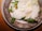 A gourmet dish featuring foamy sauce, delicate greens, and a small yellow flower arranged on a rustic ceramic plate. The presentation is artistic and elegant.