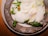 A gourmet dish featuring foamy sauce, delicate greens, and a small yellow flower arranged on a rustic ceramic plate. The presentation is artistic and elegant.