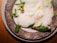 A gourmet dish featuring foamy sauce, delicate greens, and a small yellow flower arranged on a rustic ceramic plate. The presentation is artistic and elegant.