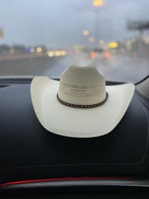 A white cowboy hat with a brown band is resting on a dark car dashboard. The background shows a blurred view of a rainy or overcast road with colorful bokeh lights from other vehicles and streetlights in the distance.