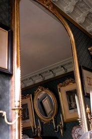A luxurious room featuring an ornate gold-framed mirror. The background includes intricately framed pictures and a detailed ceiling with elaborate moldings. Candlestick wall sconces are also visible, adding to the elegant decor.
