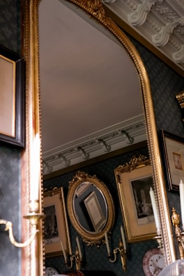 A luxurious room featuring an ornate gold-framed mirror. The background includes intricately framed pictures and a detailed ceiling with elaborate moldings. Candlestick wall sconces are also visible, adding to the elegant decor.