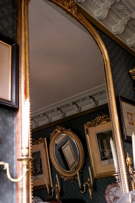 A luxurious room featuring an ornate gold-framed mirror. The background includes intricately framed pictures and a detailed ceiling with elaborate moldings. Candlestick wall sconces are also visible, adding to the elegant decor.