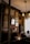 An elegant and dimly lit room features vintage decor, including ornate frames on the walls and an antique chandelier hanging from the ceiling. The room has a classic design with rich wooden furniture and warm tones. A decorative fireplace mantel displays a small sculpture, while a series of portraits or paintings adorn the walls. Soft light filters through curtains, creating a cozy and historical atmosphere.
