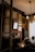 An elegant and dimly lit room features vintage decor, including ornate frames on the walls and an antique chandelier hanging from the ceiling. The room has a classic design with rich wooden furniture and warm tones. A decorative fireplace mantel displays a small sculpture, while a series of portraits or paintings adorn the walls. Soft light filters through curtains, creating a cozy and historical atmosphere.