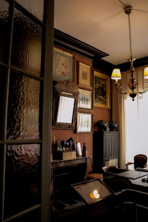 An elegant and dimly lit room features vintage decor, including ornate frames on the walls and an antique chandelier hanging from the ceiling. The room has a classic design with rich wooden furniture and warm tones. A decorative fireplace mantel displays a small sculpture, while a series of portraits or paintings adorn the walls. Soft light filters through curtains, creating a cozy and historical atmosphere.