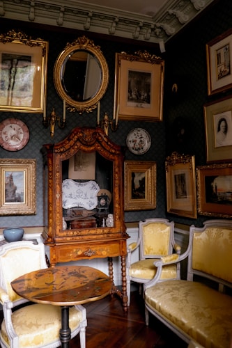 A quaint, richly decorated room with ornate wooden furniture and a collection of framed artworks and mirrors on the walls. The room features a central wooden cabinet with intricate carvings, flanked by upholstered chairs with a yellow floral pattern. The mirror above the cabinet reflects light into the room, creating an atmosphere of warmth and elegance. Dark wood floors and deep green wallpaper add to the vintage aesthetic.