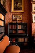 An elegant, vintage library room features ornate wallpaper and framed paintings on the walls. A dark wood bookshelf filled with various books sits against the wall, with a decorative globe on top. A plush, upholstered chair with rich orange fabric is placed nearby, creating a warm and sophisticated atmosphere.