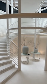 A modern, minimalist interior space characterized by curved white staircases and railings. Two white tables with large vases containing bare branches are placed on the floor, adding to the simplistic decor. The setting is serene and well-lit, emphasizing clean lines and neutral tones.