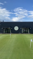 A well-maintained putting green with several small flags stands in front of a modern, dark-colored building. The building features large glass doors and a circular sign above one of the entrances, indicating it is part of a golf club. The sky is clear with scattered clouds.