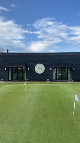 A well-maintained putting green with several small flags stands in front of a modern, dark-colored building. The building features large glass doors and a circular sign above one of the entrances, indicating it is part of a golf club. The sky is clear with scattered clouds.