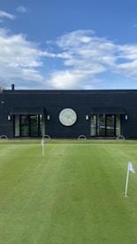 A well-maintained putting green with several small flags stands in front of a modern, dark-colored building. The building features large glass doors and a circular sign above one of the entrances, indicating it is part of a golf club. The sky is clear with scattered clouds.