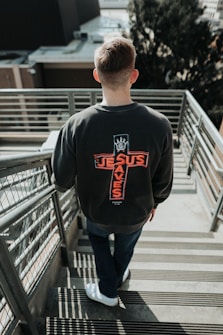 A person with short hair descends an outdoor metal staircase. They are wearing a dark sweatshirt with the phrase 'Jesus Saves' printed in red on the back, arranged in the shape of a cross. The surrounding area includes buildings and blurred trees in the background.