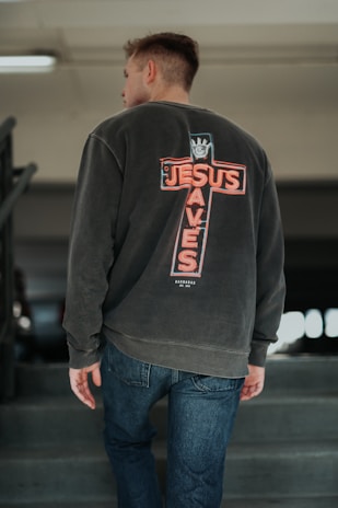 A person is walking up a concrete staircase wearing a dark sweatshirt with a design on the back that resembles a neon cross with the words 'JESUS SAVES'. They are also wearing blue jeans. The background is blurry, possibly indicating an indoor setting, with metal railings on the side.