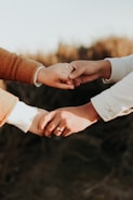 Two hands are gently holding each other, with one hand clasped onto a finger. The hands are wearing rings, indicating a possible connection or relationship, and they are positioned against a blurred, natural background.