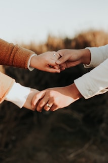 Two hands are gently holding each other, with one hand clasped onto a finger. The hands are wearing rings, indicating a possible connection or relationship, and they are positioned against a blurred, natural background.