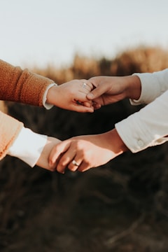 Two hands are gently holding each other, with one hand clasped onto a finger. The hands are wearing rings, indicating a possible connection or relationship, and they are positioned against a blurred, natural background.