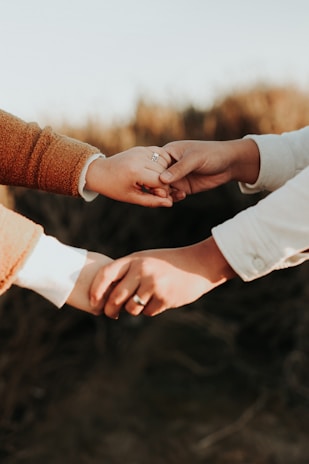 Two hands are gently holding each other, with one hand clasped onto a finger. The hands are wearing rings, indicating a possible connection or relationship, and they are positioned against a blurred, natural background.