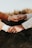 Two hands are gently holding each other, with one hand clasped onto a finger. The hands are wearing rings, indicating a possible connection or relationship, and they are positioned against a blurred, natural background.