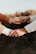 Two hands are gently holding each other, with one hand clasped onto a finger. The hands are wearing rings, indicating a possible connection or relationship, and they are positioned against a blurred, natural background.