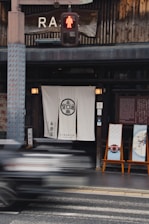 A street scene featuring the exterior of a traditional Japanese store with a white curtain displaying Japanese characters at the entrance. Two decorative panels with Japanese art are placed on easels outside. Above, there is a large wooden sign with partial text, and a pedestrian crossing signal is lit with a red figure. A blurred vehicle is passing by in the foreground, suggesting motion.