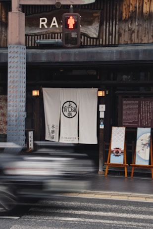 A street scene featuring the exterior of a traditional Japanese store with a white curtain displaying Japanese characters at the entrance. Two decorative panels with Japanese art are placed on easels outside. Above, there is a large wooden sign with partial text, and a pedestrian crossing signal is lit with a red figure. A blurred vehicle is passing by in the foreground, suggesting motion.
