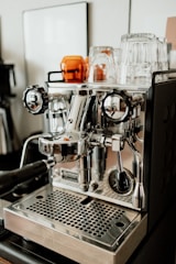 A shiny, stainless steel espresso machine with various knobs and levers is set on a counter. Several clear and amber glass cups are placed on top of the machine. The environment appears to be a modern kitchen or a cafe setting.
