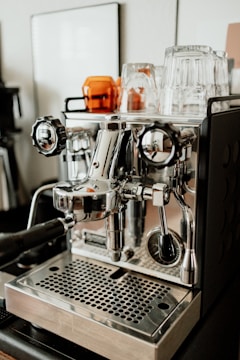 A shiny, stainless steel espresso machine with various knobs and levers is set on a counter. Several clear and amber glass cups are placed on top of the machine. The environment appears to be a modern kitchen or a cafe setting.